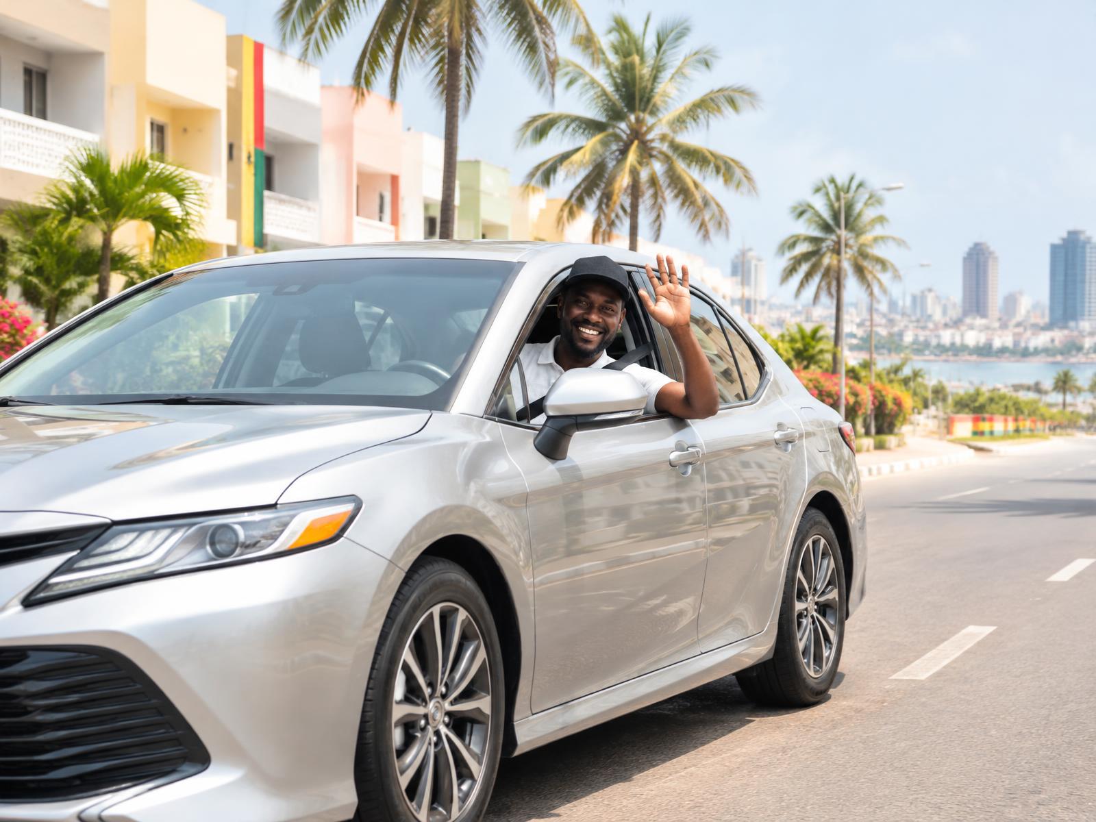 A smiling ChaleRide driver waving from a silver sedan on a sunny Accra street